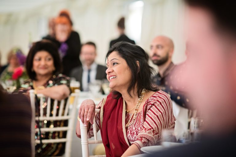 Mother of the bride listens to the father of the brides wedding speech. Mother of the bride in focus. Other people in the picture out of focus. Photographed at North Mymms Park, by Blooming Photography.