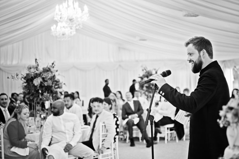 Groom stands in the marquee with the guests in the background (out of focus) as he delivers his speech. (B&w photo) Photographed at North Mymms Park, by Blooming Photography.