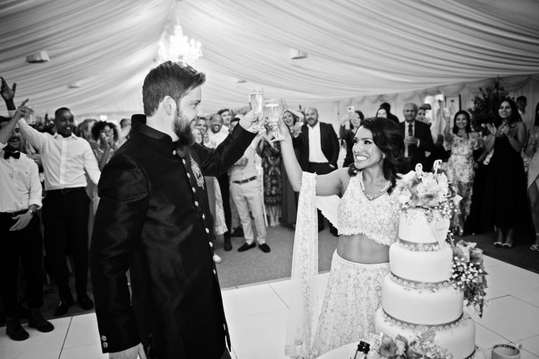 Bride and groom raise their glass and cheers each other as guests behind them (out of focus) cheer too. They are standing next to the impressive wedding cake. Black and white photo. Photographed at North Mymms Park, by Blooming Photography.