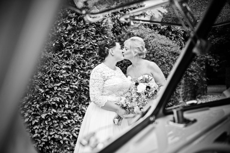 B&W candid photo of bride and bride kissing, shot through the window of a old VW campervan.