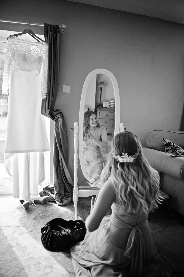 B&W portriat image of bridesmaid doing her hair in the mirror with the brides wedding dress hung up.
