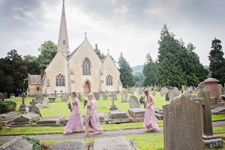 Bridesmaids walk up the path with the iconic Leckhampton, St Peters Church in the foreground.