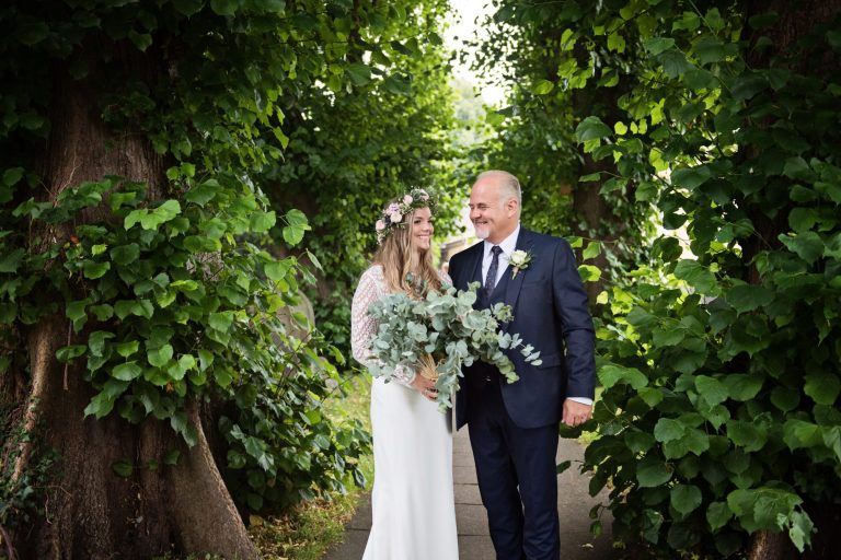 Bride and her father together, smiling amongst tall lime trees and green leaves. Photo by Blooming Photography.
