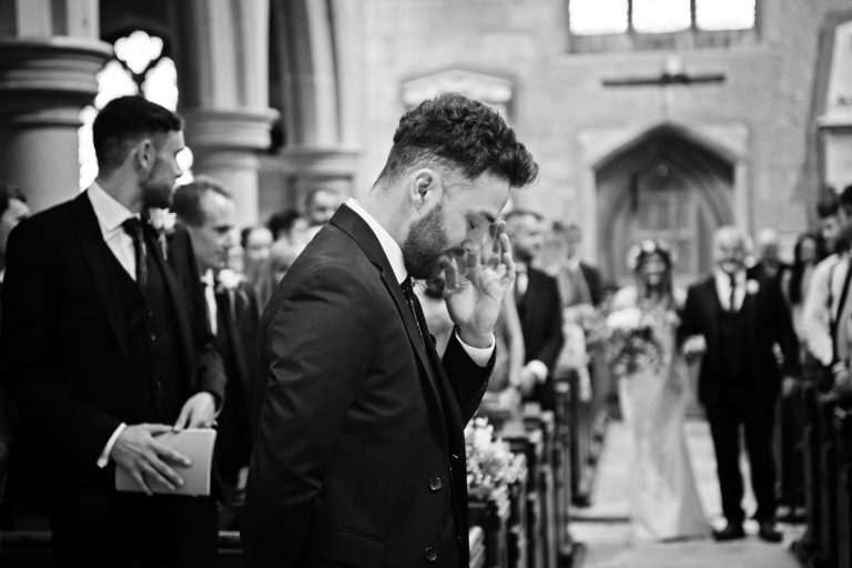 B&W photo of focused on the groom as he wipes a tear away from his eye as the bride walks down the wedding isle towards him with the brides dad proudly holding her arm.