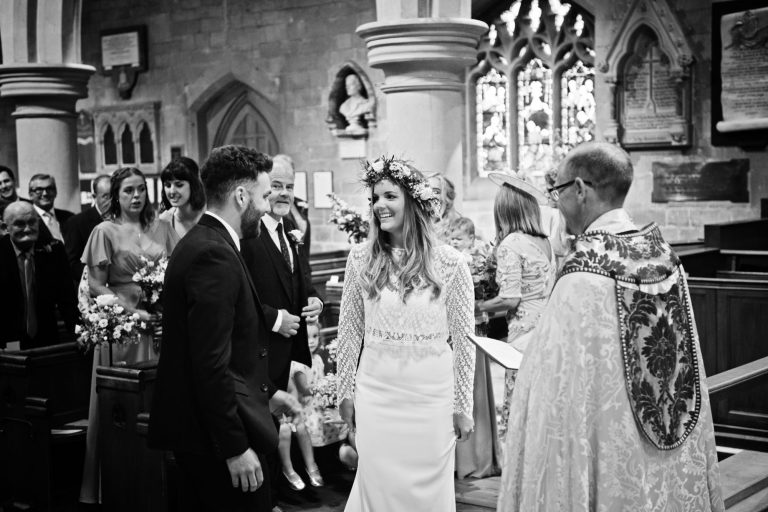 B&W candid photo of Overview of the bride and groom saying their wedding vows with the congregation aka wedding guests stood behind them.