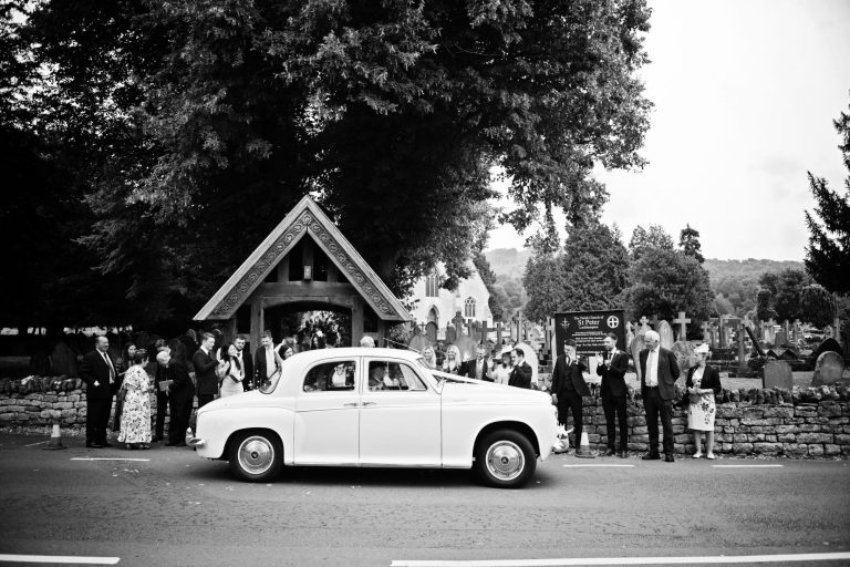 B&W photo looks like from the 1950's outside St Pauls's Church, Leckhampton. Guests waiving goodbye to the bride and groom as they leave in their classic car.