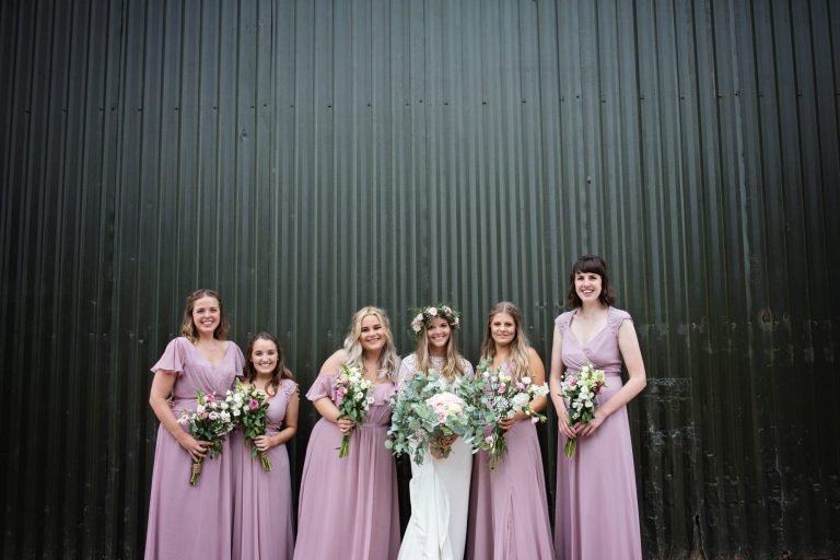 Bride and bridesmaids pose with their wedding flower bouquets pose in front of an agricultural building.