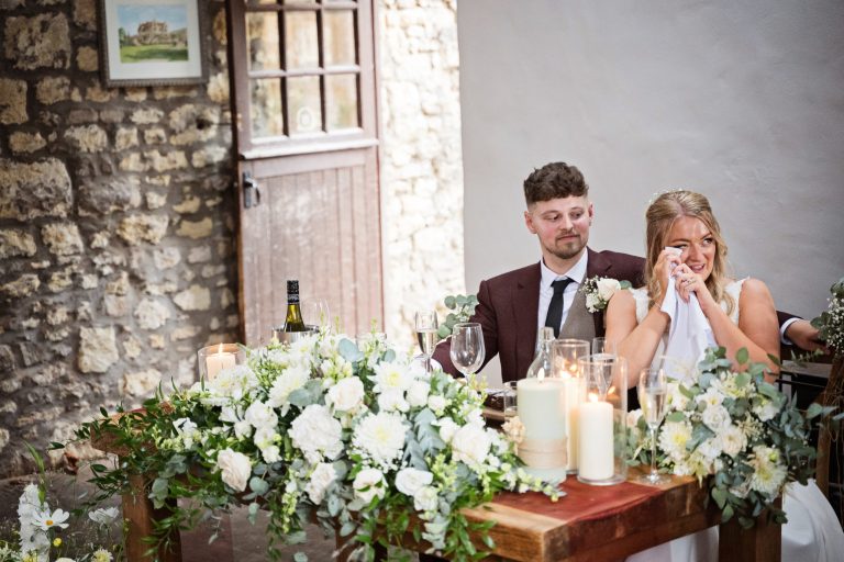 a bride and groom sat at their wedding breakfast table (surrounded by candles and flowers). The bride using a napkin to wipe her eye during an emotional speech.