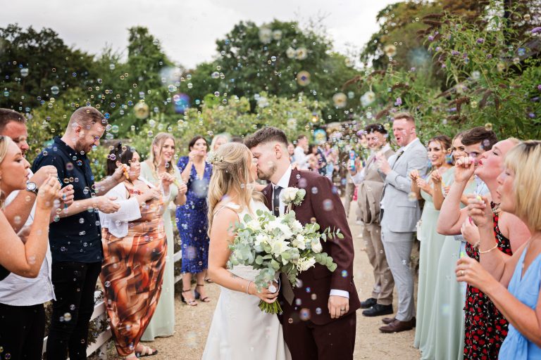 Bride & Groom kissing whilst their guests are celebrating by blowing bubbles behind them.