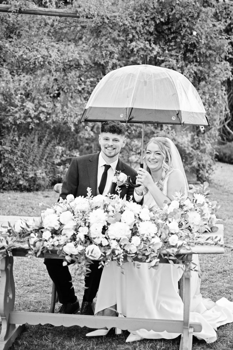 B&w image of a bride and groom smiling whilst holding an umbrella as they've just signed their wedding register.