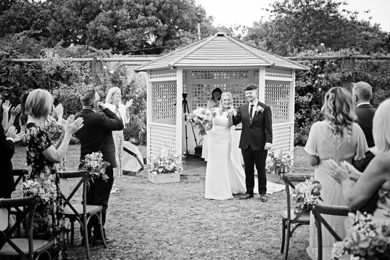 B & W image of bride and groom smiling with their friends and family clapping them.