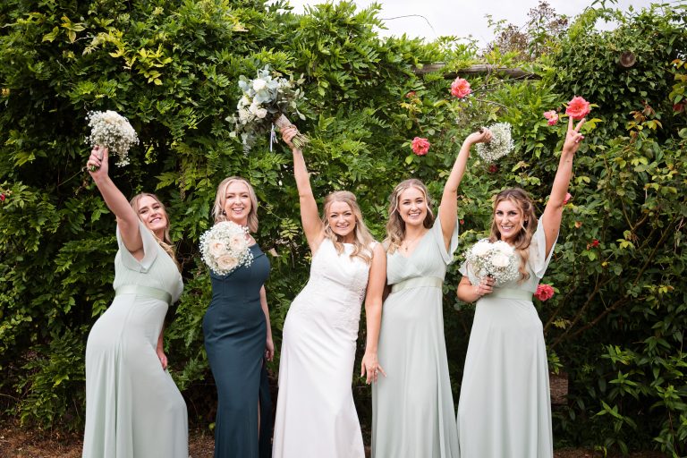 bridesmaid and bride stand against a floral rose wall with their hands in their air (holding their flower bouquets), smiling
