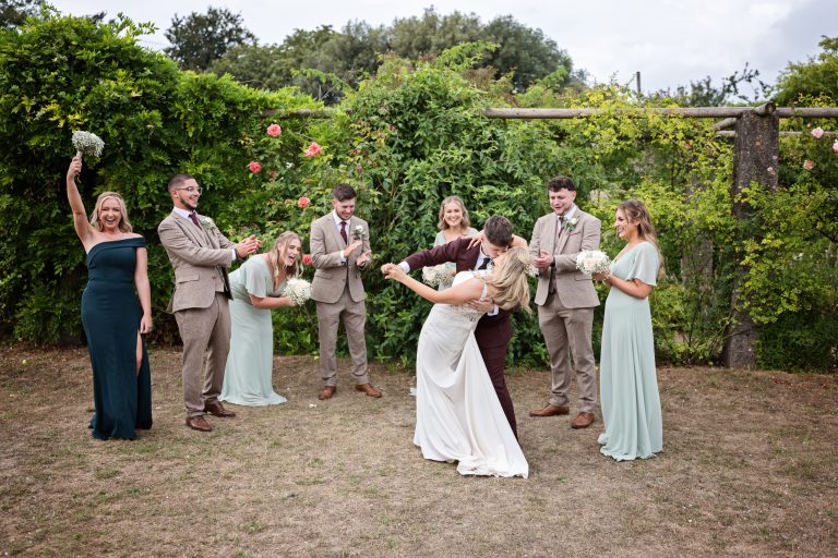 Bride and groom dip and kiss in a garden with their bridal party celebrating them
