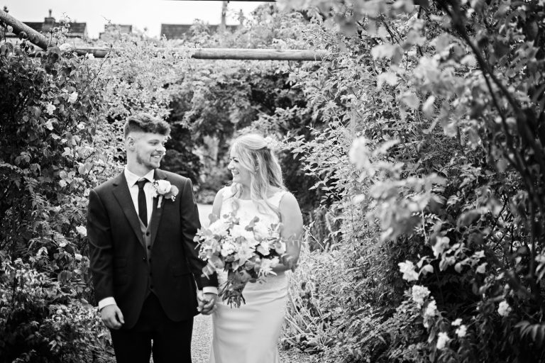 b&w image of bride and groom smiling at each other as they walk through a rose garden.