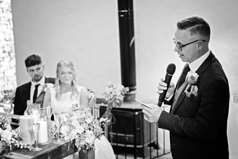 B&w image of father of the groom doing a speech. The bride & groom are sat behind him.