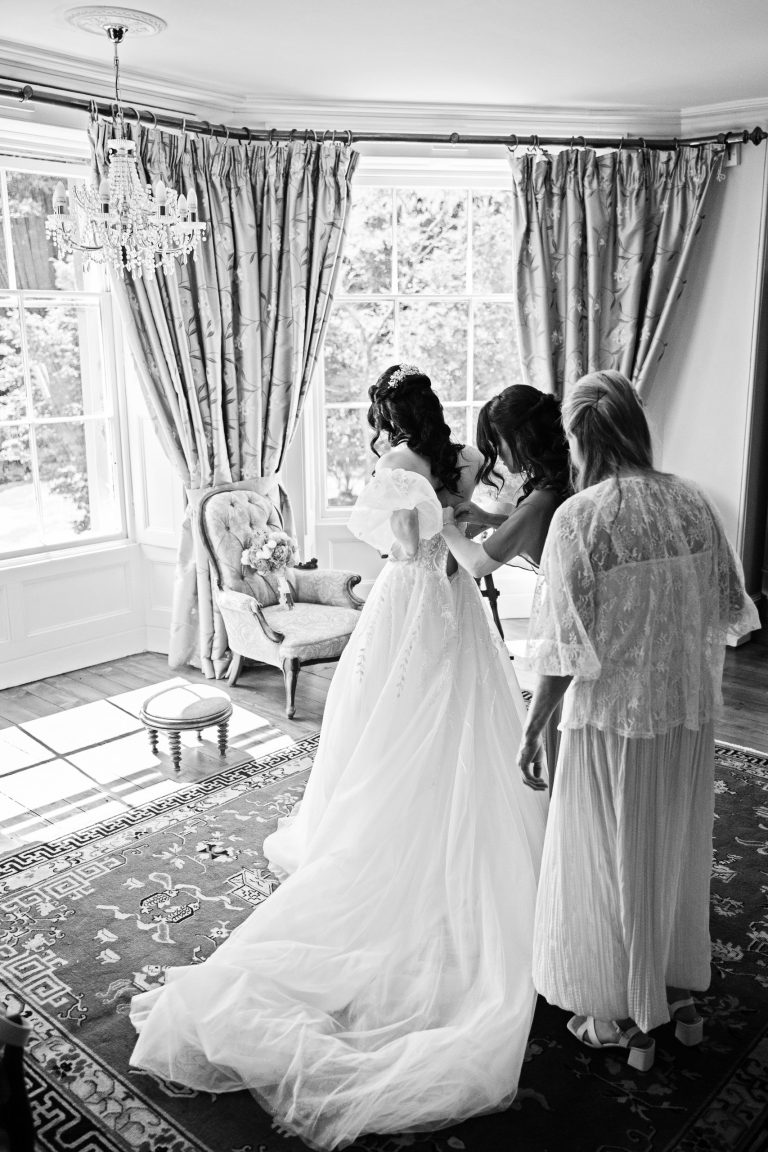 A B&W image of a bride bride getting in her wedding dress - in front of a gorgeous bay window