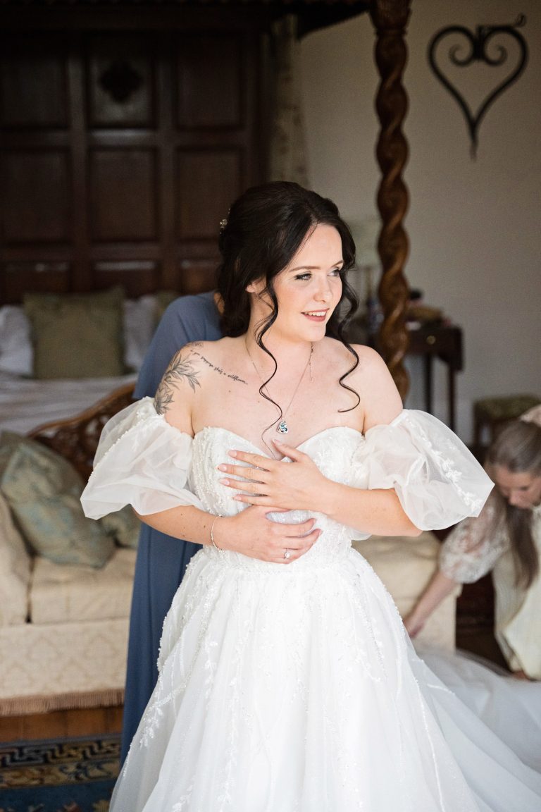 A portrait image of a bride getting into her stunning wedding dress, mother of the bride can just be seen to be laying out the dress