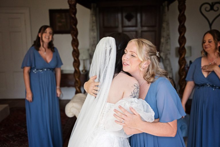 A bridesmaid giving the bride a hug with other bridesmaids in blue dresses and a four poster bed behind her