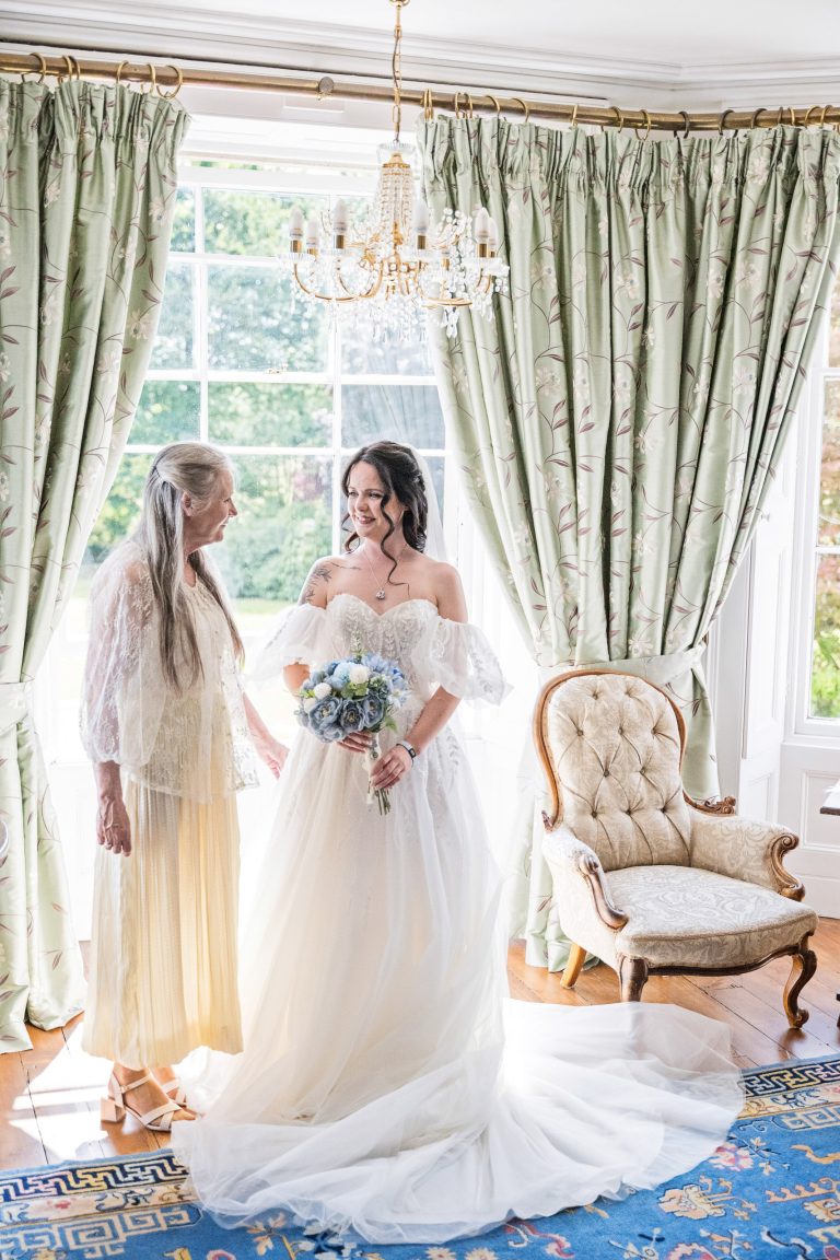A bride and her mother stand together in front of a large bay window.