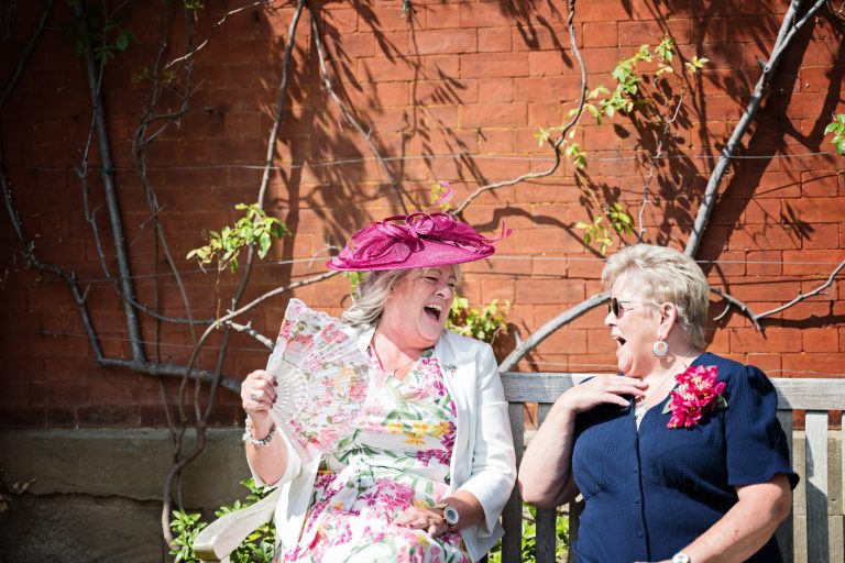 two ladies sitting on a bench, laughing at each other, one lady has a pink fascinator and is fanning her self as it is clearly a hot day