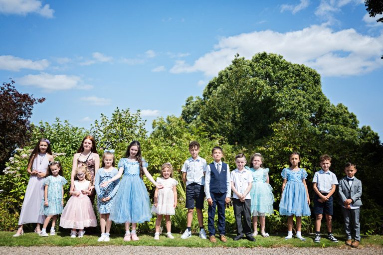 A row of children line up smiling with large shrubs behind them and a blue sky