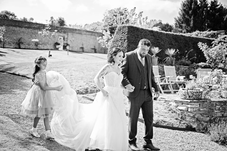 Unscripted candid wedding photo of a bride and groom walking holding hands whilst their daughter (flower girl) is carrying the train of her mums (bride) dress