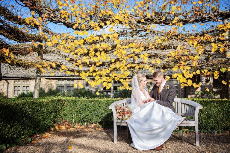 Candid photo of bride and groom holding hands and chatting as they sit on a garden bench, with autumnal pleached tree's behind them.