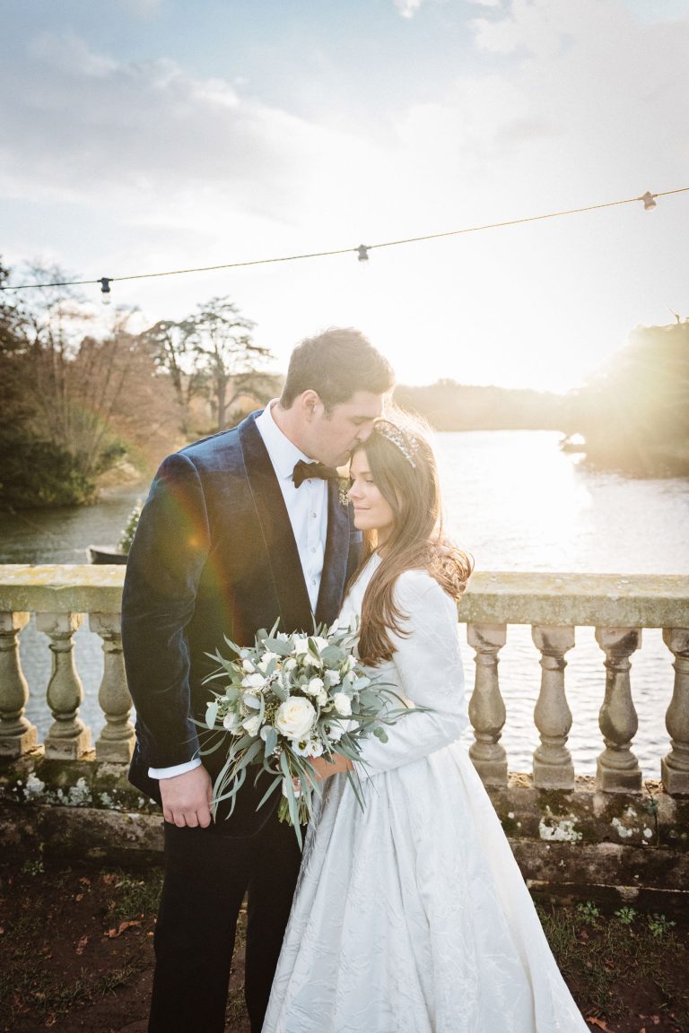 Stunning portrait photo of a bride and groom on the bridge by the lake at Compton Verney - shot at winter with a low sun in the background.