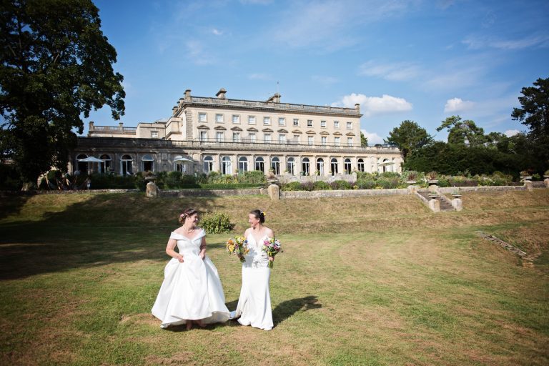 Bride and bride walk hand in hand across the lawn in front of Cowley Manor.
