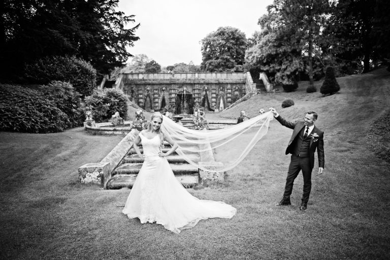 Groom lifting the brides long wedding veil up in the air in the gardens.