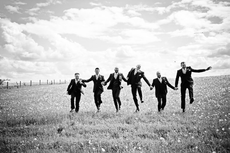 Groomsmen running towards the camera skipping and jumping in a field whilst holding hands and smiling. Fun b&w image.