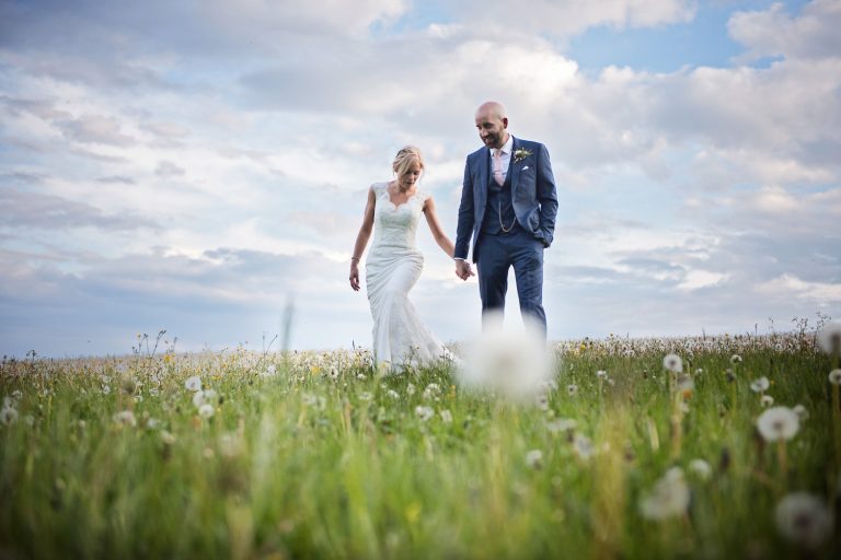 Bride and groom hold hands as they walk through a field of dandelions.