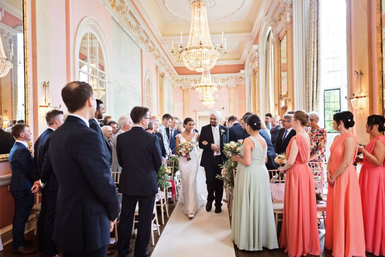 A bride walks down the isle with her father. Groom and everyone looks onward. Chandeliers and ornate fixings make this look luxurious