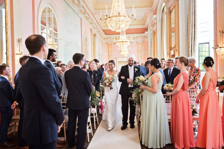 A bride walks down the isle with her father. Groom and everyone looks onward. Chandeliers and ornate fixings make this look luxurious