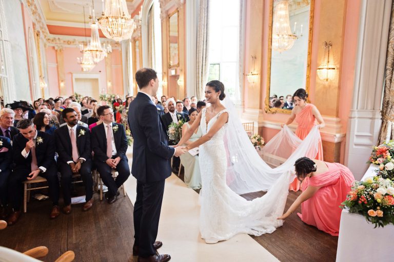 A bride greets her husband to be during their wedding ceremony at Danesfield House. Bridesmaids are tidying up the brides veil and wedding dress. Chandeliers and ornate fixings make this look luxurious