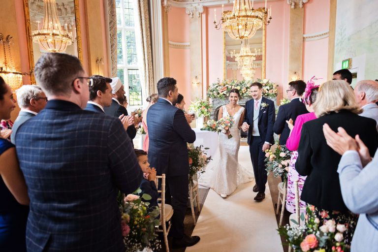 Bride and groom hold hands as their friends clap as they walk down the wedding isle after being announced husband and wife. Chandeliers and ornate fixings make this look luxurious