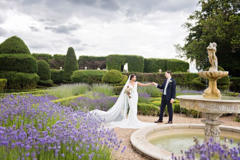 Bride and groom stand in the impressive grounds at Danesfield House. Lavender and a water fountain in front of them.