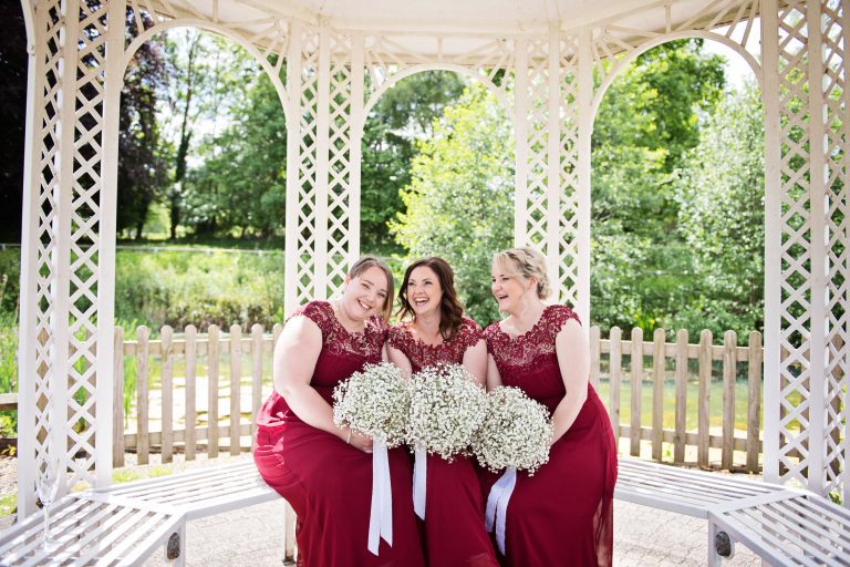 Bridesmaids dressed in red, smiling and laughing at each other under a pergola.
