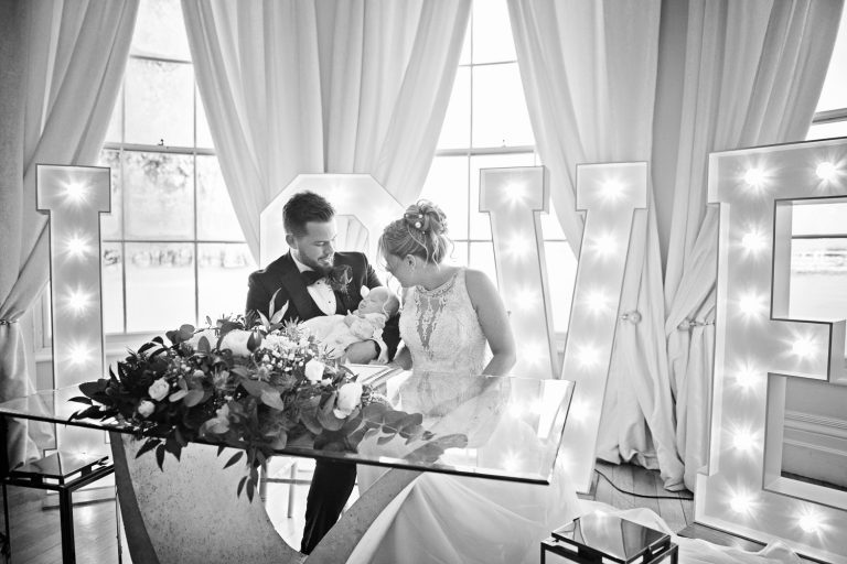 B&W image of bride and groom holding their baby in front of LOVE light up letters.