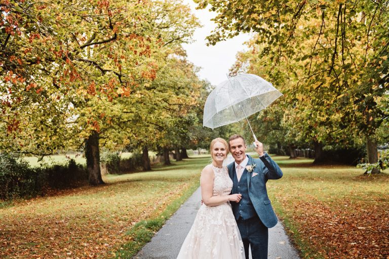 Autumnal image (lots of leaves on the floor), with a bride and groom (holding an umbrella)