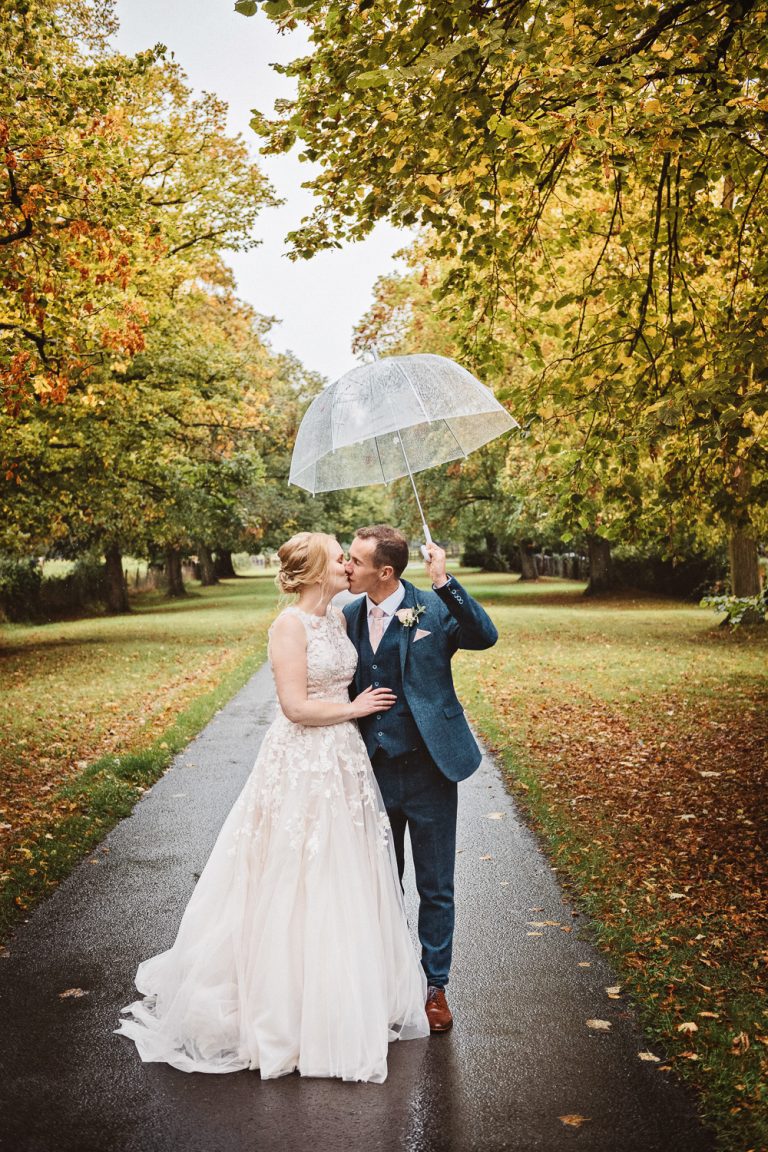 Autumnal image (lots of leaves on the floor), with a bride and groom (holding an umbrella)