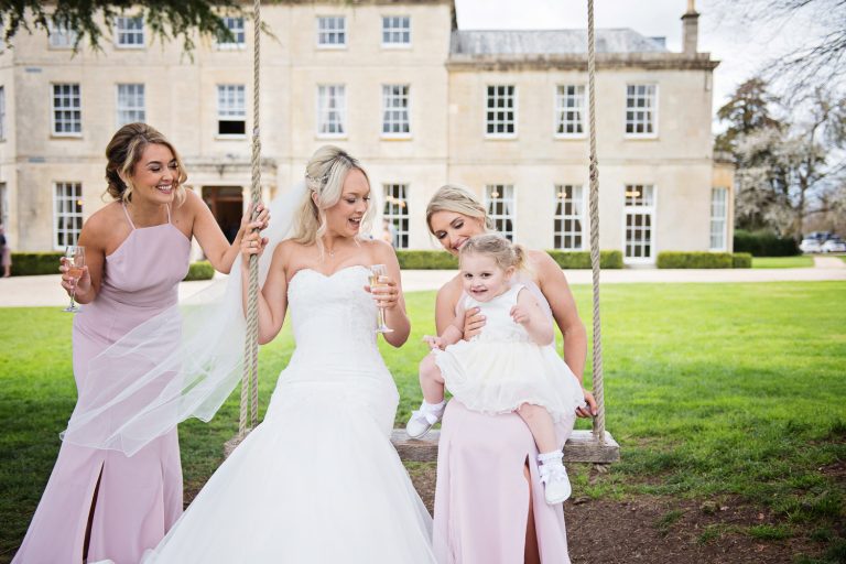 Bridesmaids, bride and flower girl sit on the swing at Eastington Park