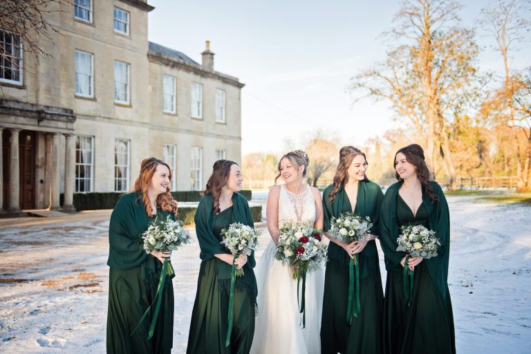 Bride and bridesmaids smiling at each other during the winter sun. Snowy backdrop