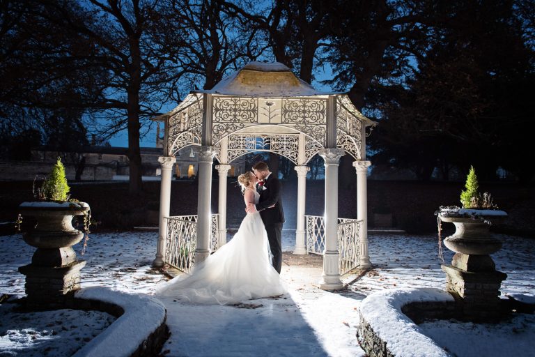 Striking night time image of bride and groom kissing under the pergola at Eastington Park with snow on the floor. Bride and groom lit up.