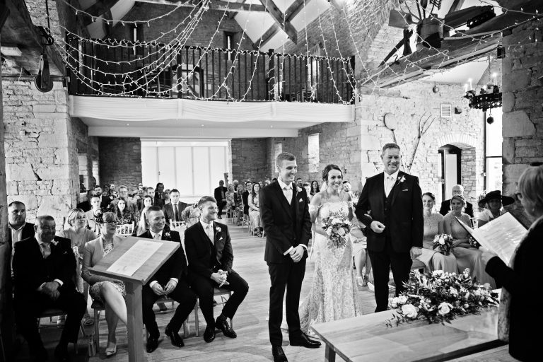 B&W photo of bride and groom and father of the bride standing at the front during their wedding ceremony at Great Tythe Barn.