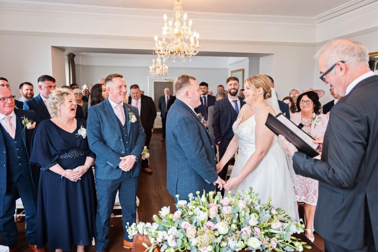 Bride and groom stand at the top of isle with their friends and family watching and smiling.