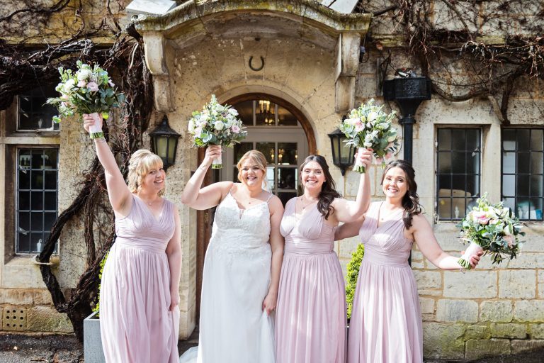 Bride and her bridesmaids raise their flower bouquets outside the stone walls of Hatton Court Hotel.