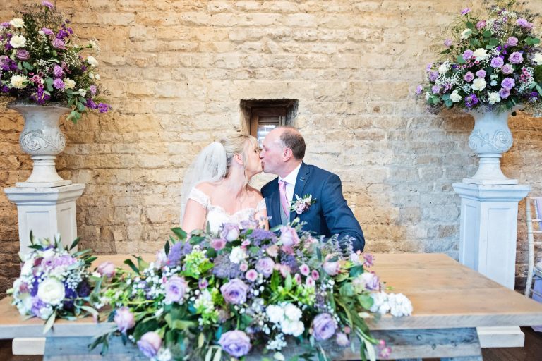 Bride and groom kiss after signing the wedding register. Gorgeous flowers in front on the table and in two urns behind them.