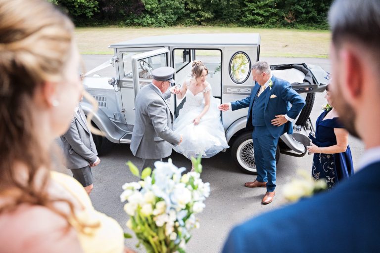 A bride gets out of Azure wedding cars with bridesmaids and family watching.