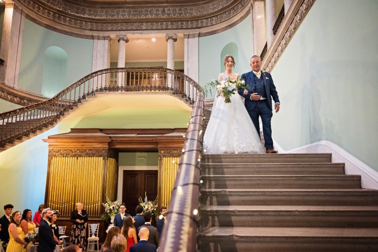 A bride walks down the stairs with her father on her way to get married. Wedding guests watching from below.